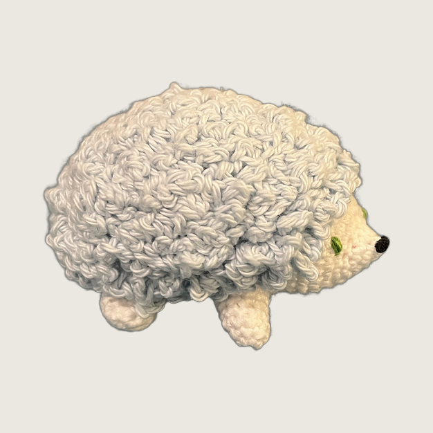 A crocheted hedgehog plush toy with a textured cream-white body and a small black nose, shown against a solid light beige background.
