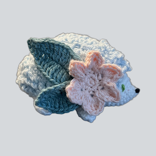 A crocheted Shaymin the Pokemon toy in white and light blue, decorated with two teal-blue leaves and a pale pink flower on its back, shown on a plain light gray background.