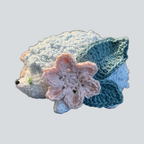 A crocheted hedgehog toy or accessory with white and light blue fur, a pink flower on its side, and teal leaf-like accents, shown on a plain light gray background.