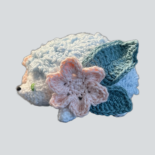 A crocheted hedgehog toy or accessory with white and light blue fur, a pink flower on its side, and teal leaf-like accents, shown on a plain light gray background.