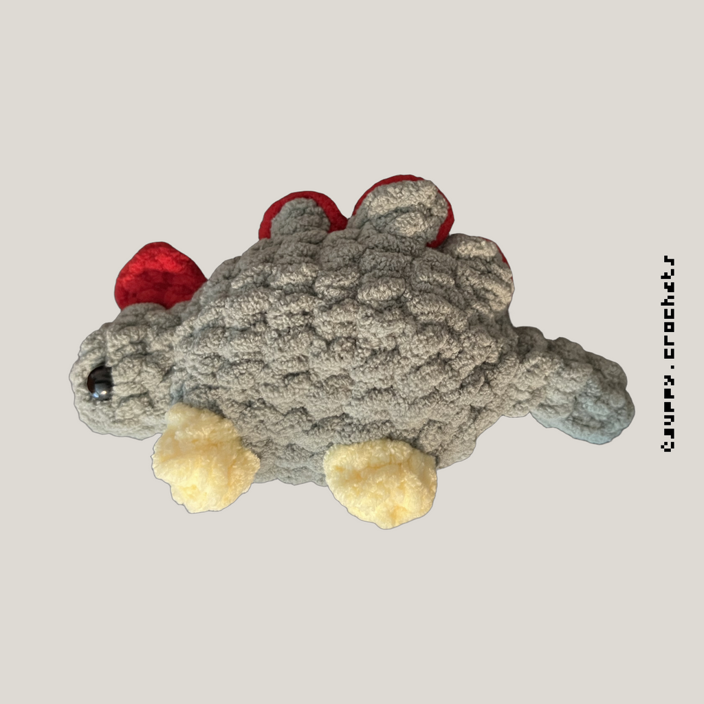 A low sew crocheted dinosaur toy in textured gray yarn with red and pale yellow accents, shown on a plain light gray background. Similar to the dinosaur featured in Stardew Valley.