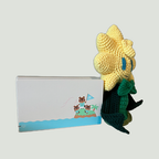 A handmade crocheted plush toy resembling a yellow flower or sun with blue eyes and a green scarf, posed next to a Nintendo switch featuring a small cartoon island illustration, on a plain light gray background.
